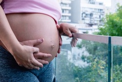 close up belly of pregnant caucasian woman with hands touching her, standing on the balcony of her house with her husband behind her, with the view towards the city and copy space.