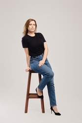 Young woman in black t shirt and jeans posing on tall wooden chair over grey background