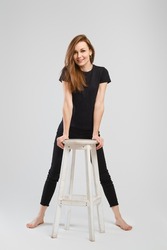 Full length studio portrait of young barefoot woman in black jean and shirt leaning to a tall chair