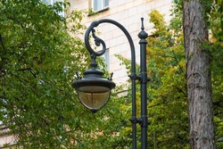 Old black cast iron lantern with round lampshade in park against background of green trees. Retro-style city landmark for street lighting.