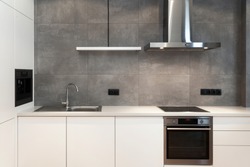 Modern interior design at white contemporary kitchen in loft style. Glossy cabinet with built in household appliance, electric stove, oven, sink on worktop and extractor hood on grey wall