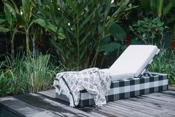 White mattress and cozy plaid on comfortable rattan deckchairs standing on wooden floor near poolside, against natural green background with copy space