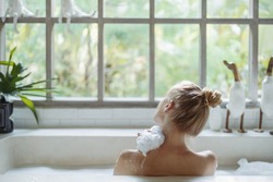 Skin care concept. Back view of young adult woman taking bath, washed shoulder, holding sponge in hand and lying in foam water, spending morning in bathroom