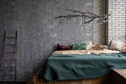 Comfortable bed with pillows, plaid and duvet in cozy bedroom under wooden branch with bulb lamp. Loft interior design and home decor in fancy apartment against grey copy space wall on background
