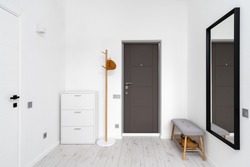 Minimalist hallway with modern interior design in contemporary apartment. Comfortable shoe storage bench near black frame mirror on white wall. Wooden coat hanger in hall with chest drawers
