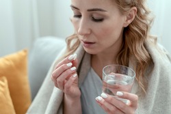 Unhealthy sick young woman covered in a plaid, takes sleeping pill from insomnia or headache. Female with glass of water, holds antidepressant meds or painkiller for menstrual pain