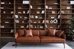 Leather sofa with cushions standing on living room with stylish interior design and collections books on bookshelves in library. Work cabinet in modern apartment