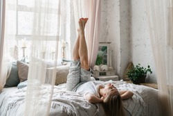 Relaxation, recreation concept. Dreamy and smiling young adult woman in pajamas raised her legs up lying on comfort bed in boho interior bedroom. Happy female spending early morning at home