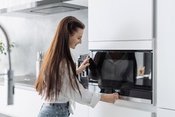 Smiling beautiful woman housewife preparing food in electric microwave oven, adjusting temperature on it. Happy female using modern kitchen built-in appliance