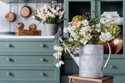 Vintage watering can filled with lots of different flowers and greenery, sitting on top of wooden ladder, comfortable kitchen enviroment, green furniture in blurred background