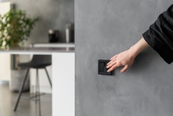 Cropped shot of woman turning light on or off in kitchen using black switch located on grey wall, modern kitchen interior design with plants, furniture and appliances in blurred background