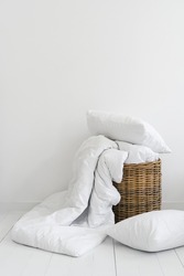 Vertical view of clean bed linen in a wicker basket. Cozy bedroom with white bedding, cotton pillow, sheets and duvet ready for laundry. Household objects in bright interior design at modern apartment