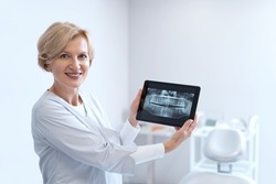 Professional dentist standing in stomatology clinic, holding digital tablet with healthy teeth x-rays, showing oral radiography on camera and smiling wide. Healthcare, medical help concept