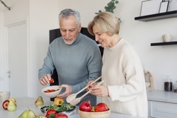 Retirement senior couple cooking healthy food together, standing on kitchen, making vegetarian salad with fresh farm vegetables. Concept of dieting and correction of eating habits