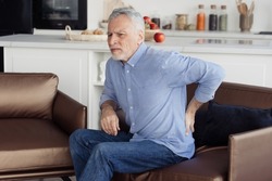 Side view photo of suffering senior man feeling lower back pain, sitting on comfortable orthopedic armchair in incorrect pose. Chronic injury, muscle inflammation and backache concept