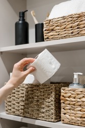 Cropped view of woman hand take toilet paper from wicker box. Domestic and household items on storage shelf in bathroom. Cosmetics bottles, clean and fresh towel in restroom closet