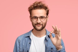 Portrait of handsome man has stubble, makes ok sign, agrees or likes something has joyful expression, poses against pink background, proves everything goes according to plan. Body language concept