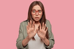 Apprehensive scared female stretches hands, refuses of something extreme, being terrified by awful proposal, asks not bother her, stares with bugged eyes through spectacles, stands indoor alone