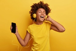 People, music, emotions concept. Delighted carefree female with Afro hairstyle dances in rhythm of melody, closes eyes listens loud song in headphones, holds smart phone has fun. Yellow color prevails