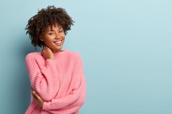 Studio shot of glad charming young female with Afro haircut, touches neck, wears oversized jumper, isolated over blue background with blank space for your promotional content. Pleasant emotions