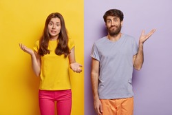 Indifferent unbothered woman and man spread hands sideways, have no idea, dressed in casual outfit, pose against different color background. Confused questioned couple with clueless expression indoor
