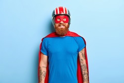 Glad bearded man with funny outlook, comes on costume party, being superhero character, wears helmet, mask and red cloak, has fun with friends, poses against blue background. Save world concept