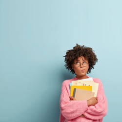 Pensive dark skinned college student holds papers and textbooks, purses lips and focused upwards, wears rosy knitted sweater, stands against blue background with copy space for your advertisement