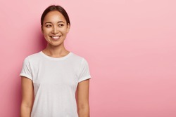 Studio shot of happy young Asian woman has tender smile, looks aside with charming expression, wears casual white t shirt, has natural beauty, isolated on pink wall. People and emotions concept