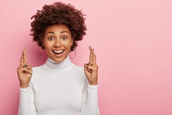 Headshot of glad smiling curly woman crosses fingers with positive expression, believes in good luck and success, looks hopefully straightly at camera, isolated over pink wall with blank space aside