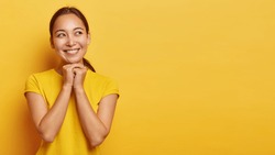 Enjoying life concept. Pleasant looking glad Asian woman looks aside, has toothy smile, keeps hands together under chin, dreams about something, wears casual clothing, isolated on yellow wall.