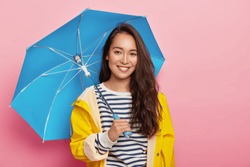 Cheerful brunette Asain woman with long dark hair, wears striped jumper, yellow raincoat, holds blue umbrella, has stroll during rainy day, isolated on pink. Bad weather doesnt spoil my mood