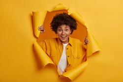 Surprised young African American woman stands in torn paper hole, dressed in stylish clothes, has excited cheerful expression, looks through breakthrough of yellow background. Wow, great news