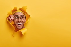 Positive male face looking curiously through paper hole, keeps hand on frame of glasses, looks aside, wears hat, sees cool advertisement or promotion, peeps with broad smile. Breaking paper.