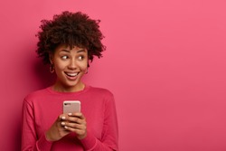 Portrait of cute curly haired girl scrolls social networks or does shopping online, looks aside positively, uses modern smartphone, wears pink sweater, poses indoor in studio, reads message.