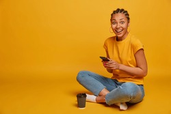 Relaxed dark skinned woman sits crossed legs, dressed in casual t shirt and jeans, uses mobile phone, sends text messages, browses internet, drinks takeaway coffee, scrolls news, isolated on yellow