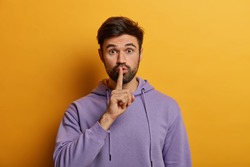 Mysterious bearded young man makes shush gesture, asks to keep secret safe, says keep voice down, quiet please, touches finger over lips, dressed in purple hoodie, isolated on yellow background