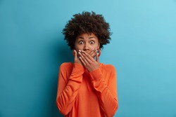 Scared shocked African American woman covers mouth, stares with eyes full of fear, dressed in casual orange jumper, cannot believe in awful news, isolated on blue background. Frightened female model