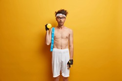 Studio shot of shocked sporty man holds dumbbell, tape measure on shoulder, wears glasses, shorts and sport gloves, stunned with his workout result, isolated on yellow background. Biceps exercise