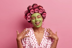 Portrait of pleased young African American woman stands with closed eyes, smiles gently, imagines something nice, applies green face mask, hair curlers, stands indoor, pink background. Domestic style