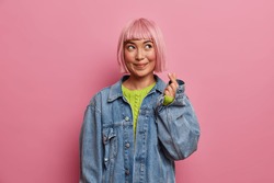 Horizontal shot of dreamy thoughtful young Asian woman makes love sign, shapes mini heart with fingers, feels happy, wears stylish denim clothes, pink background. Body language, korean style