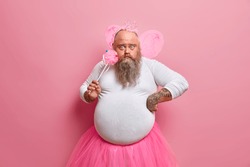 Obese bearded man dresses for hallooween party, plays fairy with magic wand, looks surprisingly at camera, wears wings and crown on bald head, being at childrens holiday. Funny dad princess.