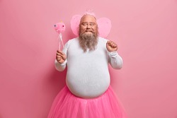 Positive plump man has fun on theme birthday party, feels like fairy who makes dreams come true, chills with children, has thick beard and fat belly, poses with magic wand and wings on back.