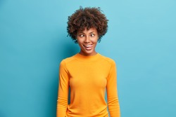 Funny crazy comic Afro American woman makes grimace and crosses eyes plays fool has fun alone sticks out tongue wears casual jumper poses against blue background. Human face expressions concept