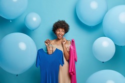 Pleased dark skinned young woman chooses stylish outfit to wear for date carries two dresses on hangers poses against blue background with inflated balloons around. People and fashion concept