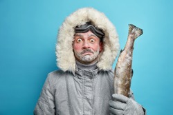 Puzzled surprised frozen man spends long hours outdoor during severe cold day dressed in grey winter jacket and gloves holds fish wears ski glasses isolated on blue background. Frosty weather