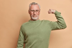 Confident satisfied grey haired man raises arm and shows muscle demonstrates results after regular training in gym wears spectacles and jumper isolated over brown background being proud of himself
