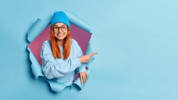 Smiling teenage girl with red hair gives recommendation or stands satisfied shows advertising content or big sale offer breaks through blue paper background. Empty space for your promotion here