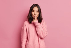 Serious mysterious brunette Asian woman presses index finger to lips makes hush gesture tells secret asks to be quiet wears long sleeved jumper poses against pink background. Secrecy concept