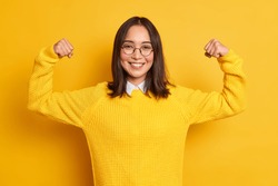 Studio shot of positive young Asian woman raises arms shows muscles pretends to be very strong and powerful smiles gently wears casual sweater isolated over yellow background. Look at my biceps