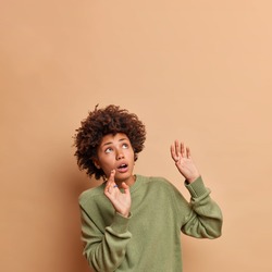 Frightened African American woman keeps arms raised tries to stop something falling above concentrated upwards being afraid wears casual jumper poses against beige background copy space area
